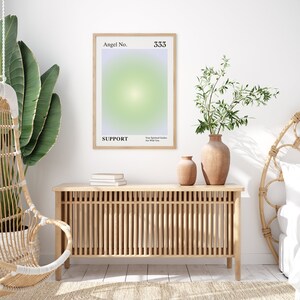 May include: A framed poster with a light green and purple gradient background. The poster has the text "Angel No. 333" and "SUPPORT" in black text. The text "Your Spiritual Guides Are With You" is also in black text. The poster is hanging on a white wall above a wooden cabinet with a slatted front.