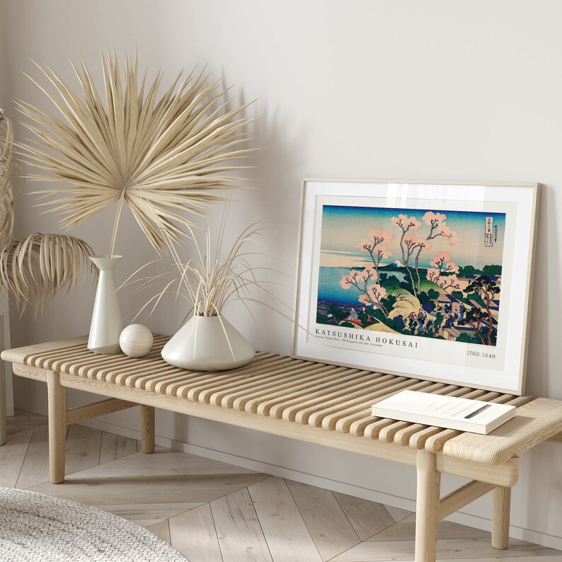 May include: A framed print of a Japanese woodblock print by Katsushika Hokusai, depicting a landscape with cherry blossoms and a view of the sea. The print is displayed on a wooden bench with a vase of dried palm leaves and a book.