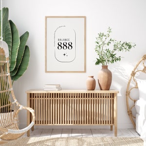 May include: A framed print with a white background and black text that reads "Everything Is Happening For a Reason, It's Meant to Be. Balance 888". The print is hanging on a white wall above a wooden cabinet with a slatted design. There are two vases with green plants on the cabinet.