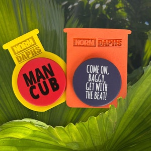 Op de afbeelding: Twee kleurrijke stickers met tekst op een achtergrond van groene bladeren. Eén sticker is geel met de tekst "NORM DAPHS" en een rode cirkel met "MAN CUB". De andere is oranje met "NORM DAPHS" en een blauwe cirkel met "COME ON, BAGGY, GET WITH THE BEAT!"