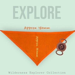Peut inclure: Un bandana triangulaire orange avec le mot "EXPLORE" en gris. Le bandana mesure environ 780 mm de long et 350 mm de large. Une étiquette en cuir avec un motif de boussole est attachée. Le texte "Wilderness Explorer Collection" est en bas.