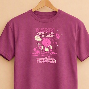 Pode incluir: Uma t-shirt de manga curta magenta escura com um gráfico de um porco cor-de-rosa vestido de piloto espacial. O texto diz HAMM SOLO e Never Tell me The Odds Kid! O design inclui uma nave espacial, um planeta e uma estação espacial.
