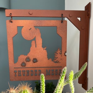 Puede incluir: Un letrero metálico de color cobre con una silueta de paisaje desértico, que incluye un sol y el texto "THUNDER MESA". El letrero está montado en un soporte y cuelga frente a una pared verde. Un cactus está en primer plano.