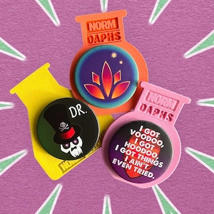 May include: Three colorful pinback buttons with unique designs and text, including a skull in a top hat, a floral design, and the phrase "I got voodoo, I got hoodoo." Each button is set against a pink background.