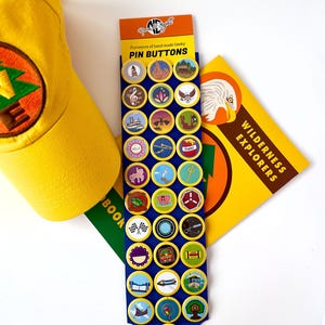 May include: A collection of colorful pin buttons with various designs, including a musical note, a teacup, and a tree. The buttons are displayed on a blue card with the text "PIN BUTTONS". A yellow hat and a book titled "WILDERNESS EXPLORERS" are also visible.
