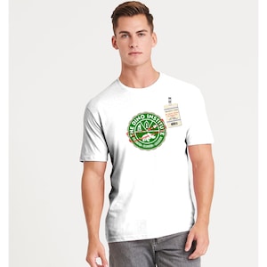 Op de afbeelding: Wit T-shirt met een groen en rood cirkelvormig ontwerp met de tekst "The Dino Institute" en een dinosaurusillustratie. Een veiligheidspas is bevestigd. Het T-shirt wordt door een persoon gedragen.