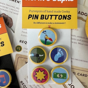 May include: A set of five round pin buttons with yellow edges and colorful designs. The designs include a bird, a telescope, a footprint, a starburst, and a dollar bill. The card reads "Norm & Daphs" and "PIN BUTTONS".