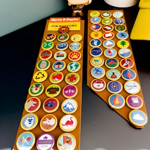 May include: A collection of colorful pin buttons with various designs, including animals, flags, and nature scenes. The buttons are arranged on brown cardboard backing. The text "Norm & Daphs PIN BUTTONS" is visible.