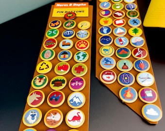 May include: A selection of colourful pin badges featuring diverse designs, such as animals, flags, and natural elements. The badges are displayed on brown card backing. The text "Norm & Daphs PIN BUTTONS" is present.