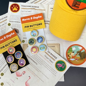May include: A yellow baseball cap with a circular patch and the letter "E", surrounded by various pin buttons with different designs, and printed cards with text and illustrations. The pin buttons are gold-rimmed.