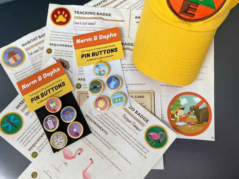 May include: A yellow baseball cap with a circular patch and the letter "E", surrounded by various pin buttons with different designs, and printed cards with text and illustrations. The pin buttons are gold-rimmed.