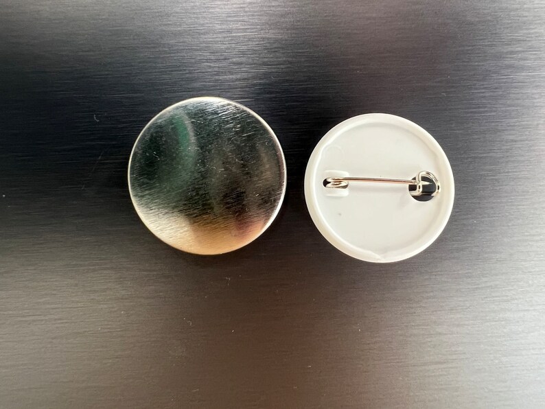 May include: Two round pinback buttons. One is silver-colored with a brushed metal finish. The other is white with a safety pin on the back. The buttons are on a dark surface.