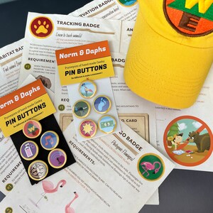 May include: A collection of pin buttons, a yellow cap, and printed cards. The pin buttons have gold rims and various designs. The cap has an embroidered patch. The cards feature text and illustrations, including a flamingo and a bear.