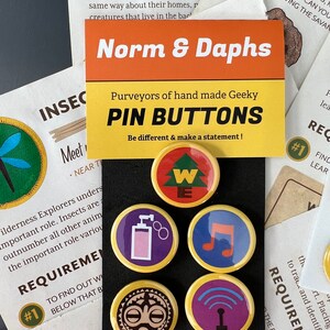 May include: A collection of five round pin buttons with gold rims. Each button features a unique design, including a tree, a perfume bottle, a musical note, a tribal mask, and a radio tower. The text "Norm & Daphs" is visible.