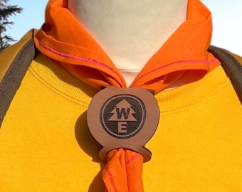 May include: A close-up of a yellow shirt with an orange neckerchief and a brown slide. The slide features a circular design with a tree and the letters "W" and "E". The neckerchief is orange with pink stitching.