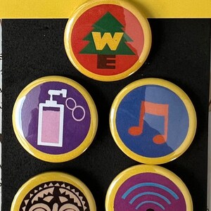 May include: A collection of five round pin buttons with yellow edges. The buttons feature various colorful designs, including a tree with the letter "W", a perfume bottle, a musical note, a tribal mask, and a Wi-Fi symbol. The text "PIN BUTTONS" and "Be different & make a statement!" are visible.