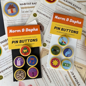 May include: A collection of colorful pin buttons with various designs, including a house, a musical note, and a paw print. The pins are displayed on black cards with the text "Norm & Daphs" and "Pin Buttons."