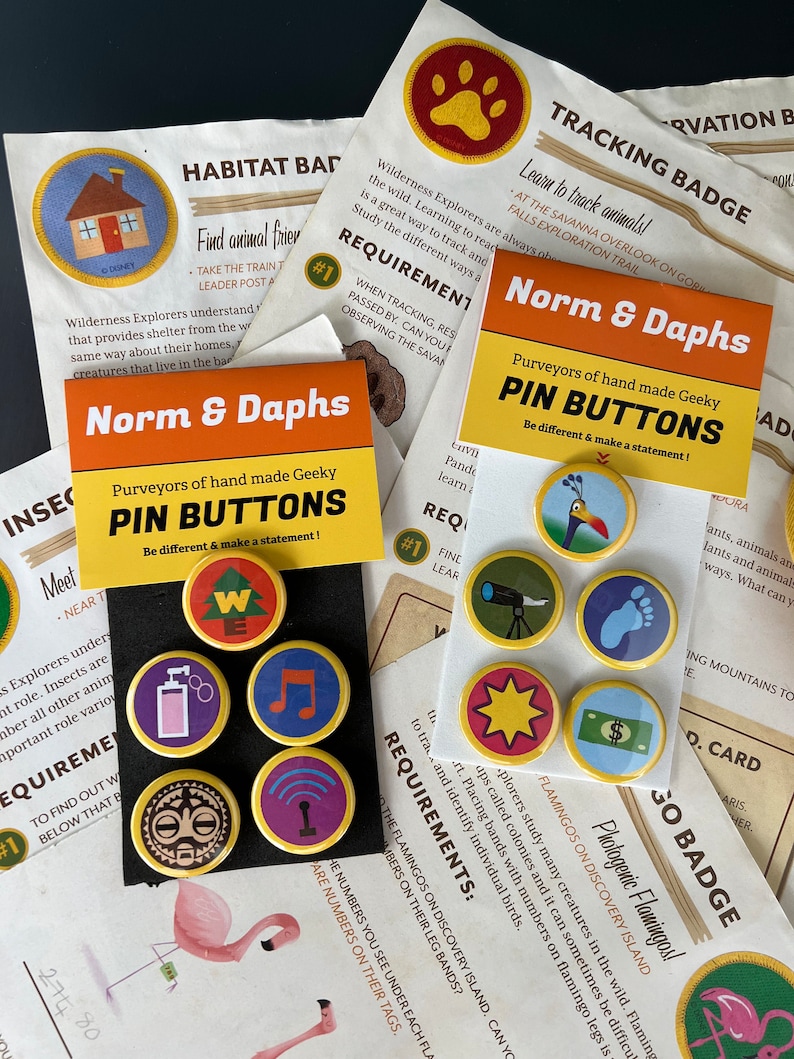 May include: A collection of colorful pin buttons with various designs, including a house, a musical note, and a paw print. The pins are displayed on black cards with the text "Norm & Daphs" and "Pin Buttons."