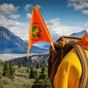 May include: An orange flag with a green and white logo is displayed against a backdrop of mountains and a blue sky. A yellow sweatshirt, orange scarf, and backpack are also visible, suggesting an outdoor theme.