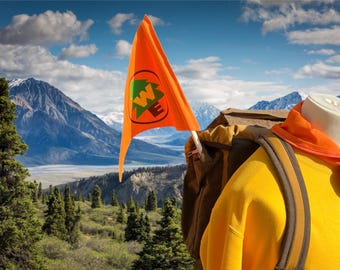 May include: An orange flag with a green and white logo is displayed against a backdrop of mountains and a blue sky. A yellow sweatshirt, orange scarf, and backpack are also visible, suggesting an outdoor theme.