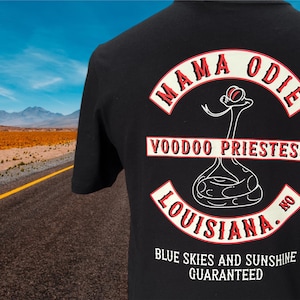 May include: Black t-shirt with a graphic design featuring the words "MAMA ODIE VOODOO PRIESTESS LOUISIANA" in a red and white arched design. The shirt also includes a snake illustration and the text "BLUE SKIES AND SUNSHINE GUARANTEED."