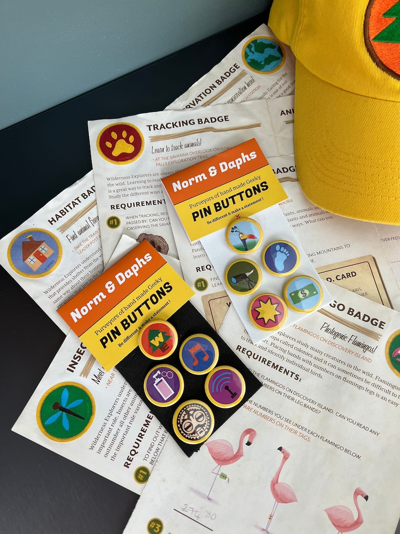 May include: A collection of colorful pin buttons with various designs, including a paw print, a footprint, and a star. The buttons are displayed with informational cards and a yellow hat with a green tree design. The text "Norm & Daphs PIN BUTTONS" is visible.