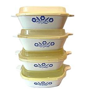 Könnte beinhalten: Ein Stapel von vier weißen Auflaufformen mit blauen Blumenmustern. Jede Form hat einen hellgelben Deckel und seitliche Griffe. Ein Vintage-Küchengeschirr-Set.