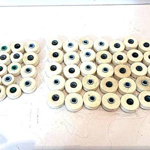 May include: A collection of small, round, off-white wooden buttons arranged in two groups. Each button features a central hole with a dark, circular insert. The buttons have a layered, disc-like appearance with visible wood grain textures.