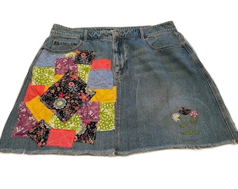 May include: A denim skirt with a patchwork design on the front. The patchwork is made up of various fabrics in different colors and patterns, including pink, yellow, green, blue, and purple. The skirt has a frayed hem and a small embroidered flower design on the right side.