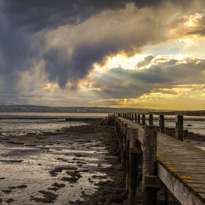 May include: A scenic view of a wooden pier stretching out over a muddy shoreline, with a dramatic sky overhead. Dark, rain-filled clouds dominate the scene, while golden sunlight bursts through the clouds on the right.