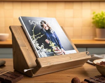 Puede incluir: Un atril de madera para libros de cocina que sostiene un libro titulado ŠOKOLADA, con una imagen de portada de una persona. El atril está rodeado de barras de chocolate negro, cacao en polvo en un cuenco de cerámica con una pala de metal y granos de cacao esparcidos.