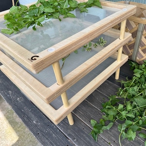 May include: A three-tiered wooden drying rack with fine mesh screens. The rack is made of light-colored wood and has a simple, functional design. Fresh green herbs are placed on the top level. The rack is designed for drying herbs and other plants.