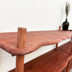 Puede incluir: Estanter&iacute;a de madera marr&oacute;n rojizo hecha a mano con un dise&ntilde;o de borde natural. La unidad presenta m&uacute;ltiples estantes sostenidos por patas cil&iacute;ndricas. Un jarr&oacute;n oscuro con plantas secas se encuentra en el estante superior. La estanter&iacute;a mide unos 152 cm de largo.