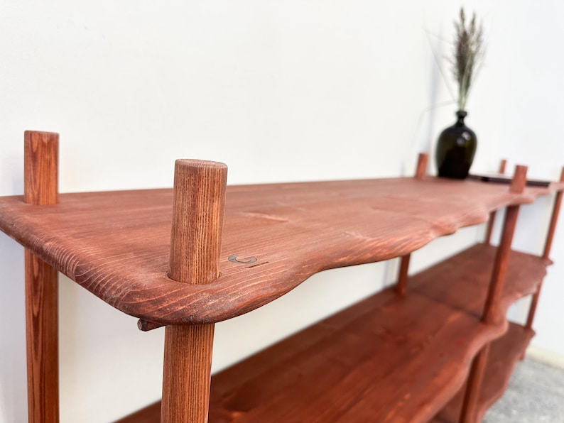 Puede incluir: Estanter&iacute;a de madera marr&oacute;n rojizo hecha a mano con un dise&ntilde;o de borde natural. La unidad presenta m&uacute;ltiples estantes sostenidos por patas cil&iacute;ndricas. Un jarr&oacute;n oscuro con plantas secas se encuentra en el estante superior. La estanter&iacute;a mide unos 152 cm de largo.