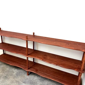 May include: A three-tiered wooden shelf unit with a rich, reddish-brown finish. The shelves have a gently undulating edge, supported by vertical posts. The unit is designed for storage or display, and is set against a white wall.