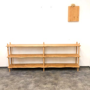 May include: A light-coloured wooden shelf unit with three shelves. The shelves have a natural, unfinished edge. The unit is supported by vertical posts and has a simple, rustic design. A small wooden plaque hangs on the wall above the shelf.