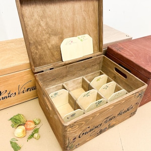 Può includere: Una scatola portaoggetti in legno marrone con coperchio incernierato e sei scomparti interni. La parte anteriore presenta la scritta "Graines Fines Herbes" in corsivo nero. Ogni scomparto contiene un piccolo divisorio chiaro con illustrazioni botaniche.
