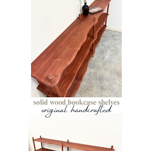 Puede incluir: Una estanter&iacute;a larga de tres niveles hecha de madera maciza con un acabado marr&oacute;n rojizo. Los estantes presentan bordes ondulados y org&aacute;nicos, sostenidos por postes cil&iacute;ndricos de madera. El texto superior dice GARKUSHA HOME y el inferior solid wood bookcase shelves original handcrafted.