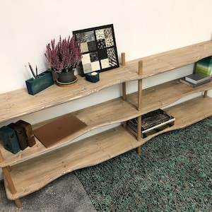May include: A three-tier wooden shelving unit with wavy, natural-toned shelves supported by vertical cylindrical posts. The unit holds a potted purple plant, a square framed art piece, a teal pencil holder, books, and a laptop on its surfaces.