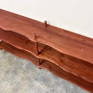 Puede incluir: Una estanter&iacute;a de madera de color marr&oacute;n rojizo de tres niveles. Los estantes tienen un dise&ntilde;o de borde ondulado y est&aacute;n soportados por clavijas verticales. La unidad est&aacute; hecha de madera te&ntilde;ida y tiene un aspecto de veta natural. El estante superior tiene una superficie plana.