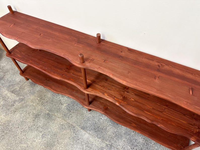 Puede incluir: Una estanter&iacute;a de madera de color marr&oacute;n rojizo de tres niveles. Los estantes tienen un dise&ntilde;o de borde ondulado y est&aacute;n soportados por clavijas verticales. La unidad est&aacute; hecha de madera te&ntilde;ida y tiene un aspecto de veta natural. El estante superior tiene una superficie plana.