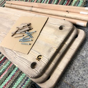 May include: A set of unfinished light-colored wooden planks with rounded corners and pre-drilled holes, accompanied by several long wooden dowels and metal screws resting on a piece of cardboard. A small black letter G logo is visible on one plank.
