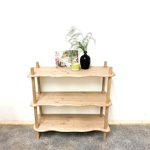 May include: A three-tiered wooden shelving unit with a natural finish. The shelves have a wavy edge design. The top shelf displays a book, a green candle, and a black vase with greenery. The unit is supported by rounded legs and vertical supports.