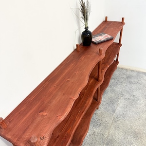 May include: A tall, multi-tiered wooden shelving unit with a reddish-brown finish and wavy, organic edges. The shelves are supported by cylindrical wooden posts at each corner. A black vase with dried grass and a small book sit on the top shelf.