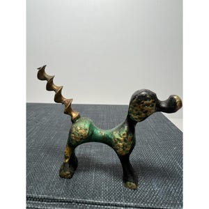 Puede incluir: Un sacacorchos de latón vintage con forma de caniche. El sacacorchos con forma de perro tiene una pátina verde y dorada, con una cola de sacacorchos en espiral. La cabeza y las patas del caniche son negras y la nariz dorada.
