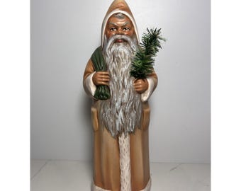 Op de afbeelding: Een staand kerstmanfiguur in een beige jas met witte bontrand en bijpassende capuchon. Het figuur heeft een lange witte baard, houdt een kleine groene zak over de schouder en draagt een miniatuur groene dennenboom in de andere hand.