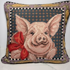 May include: A decorative pillow featuring a pink pig with a red bow. The pillow has a purple background with a black and white checked border. The pig is looking at the viewer with a friendly expression.