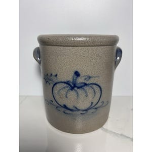 Pode incluir: Um pote de grés cilíndrico com acabamento bege salpicado e duas alças laterais. A parte frontal apresenta um desenho de abóbora azul pintado à mão com detalhes decorativos. A borda é espessa e arredondada, conferindo uma estrutura robusta.