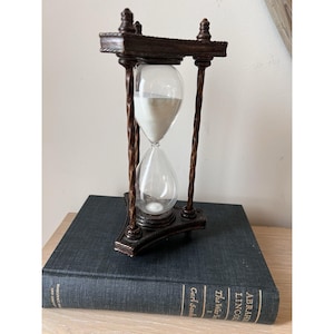 May include: A dark brown hourglass with a clear glass center filled with white sand. The hourglass has a decorative wooden frame with twisted columns and sits atop a dark blue book.
