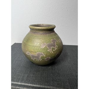 May include: Green ceramic vase with a round body and a narrow neck. The vase features a design of running horses in a lighter green colour. The vase has a textured surface and a dark brown rim.